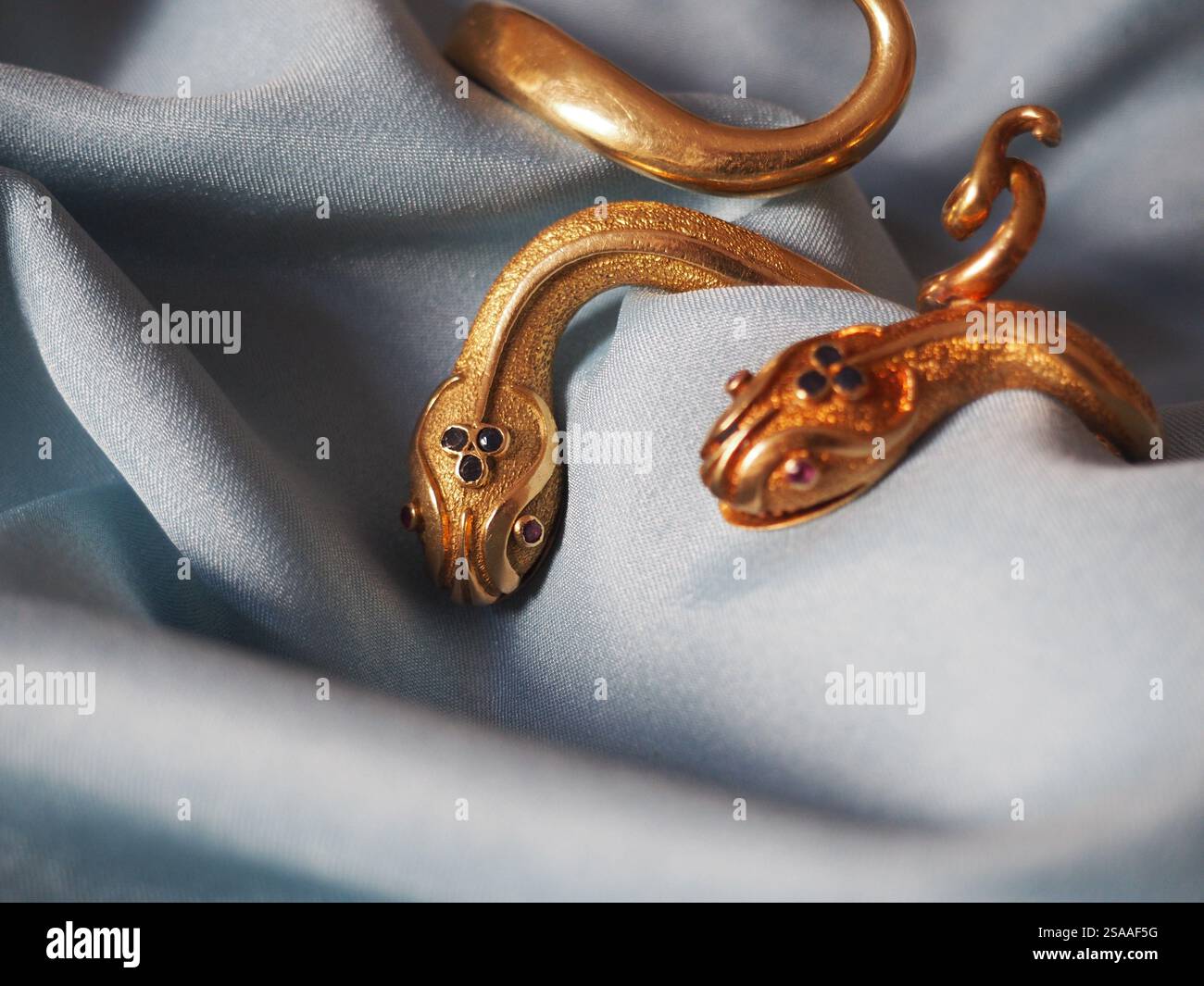 Armreifen mit goldenem Schlangenmotiv, die anmutig auf einem seidenblauen drapierten Stoff ruhen und ihr kompliziertes Design und ihre künstlerische Handwerkskunst hervorheben. Stockfoto