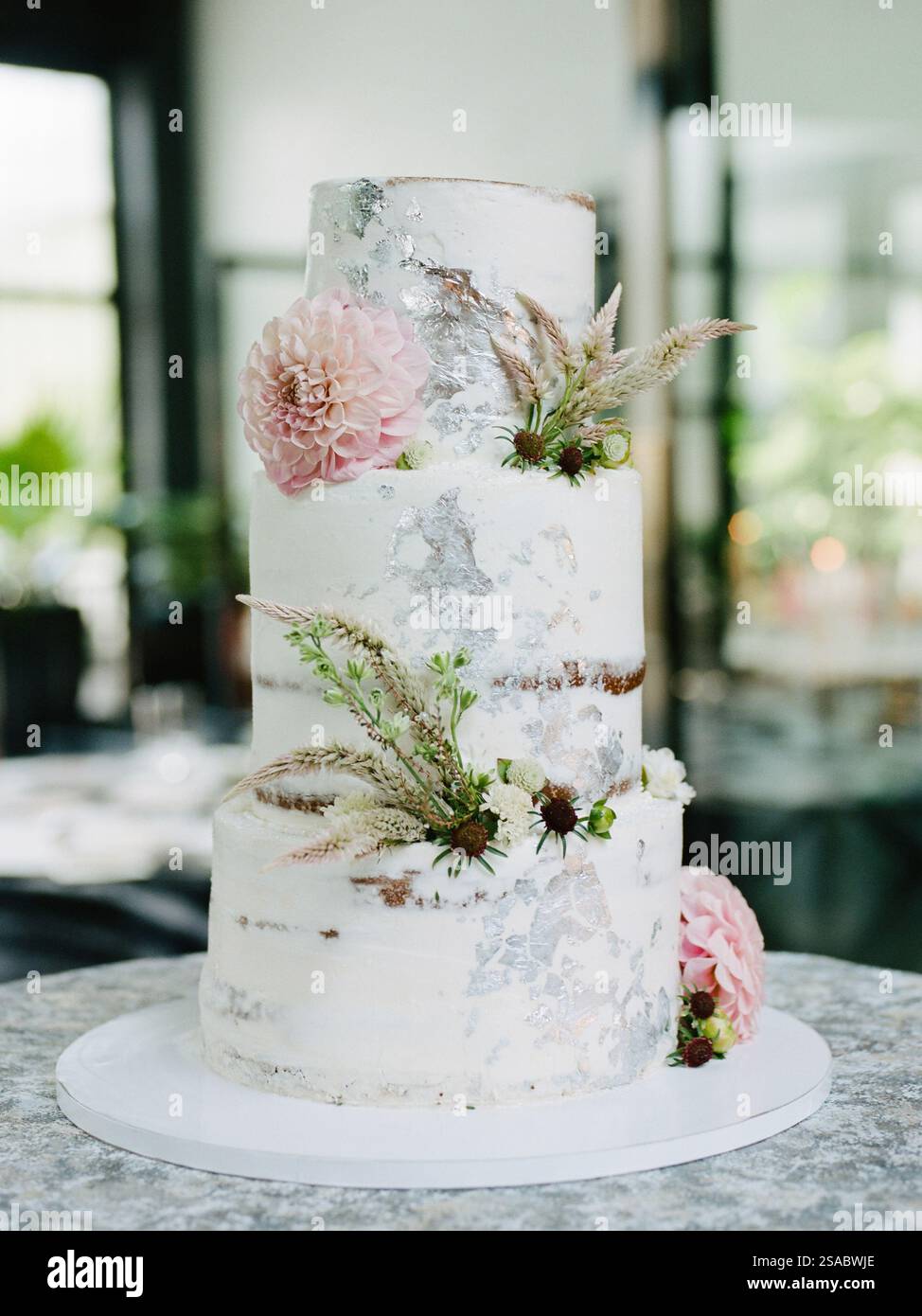 Ein wunderschön gefertigter dreistöckiger Kuchen mit glattem weißem Finish, dekoriert mit frischen Blumen und Grün, in einem schicken, modernen Ambiente voller wi-fi Stockfoto