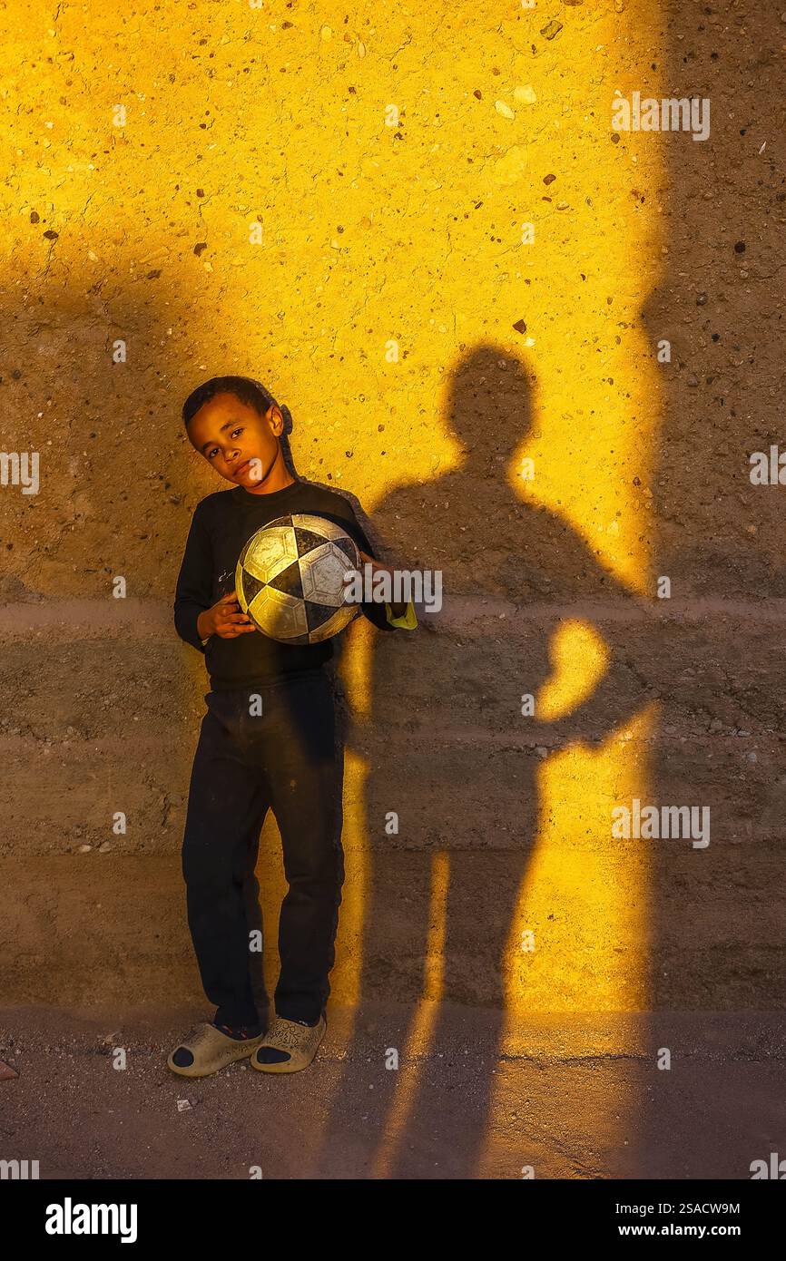 Junge, der an einer Wand steht und einen Ball hält, neben dem Schatten eines anderen Jungen, Tazert, Marokko Stockfoto