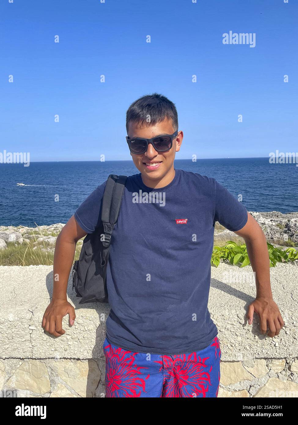 Junger Mann, der an einer Wand steht und in die Kamera lächelt, Meer im Hintergrund, Italien Stockfoto Junger Mann, der an einer Wand steht und in die Kamera lächelt, Meer im Hintergrund, Italien Stockfoto