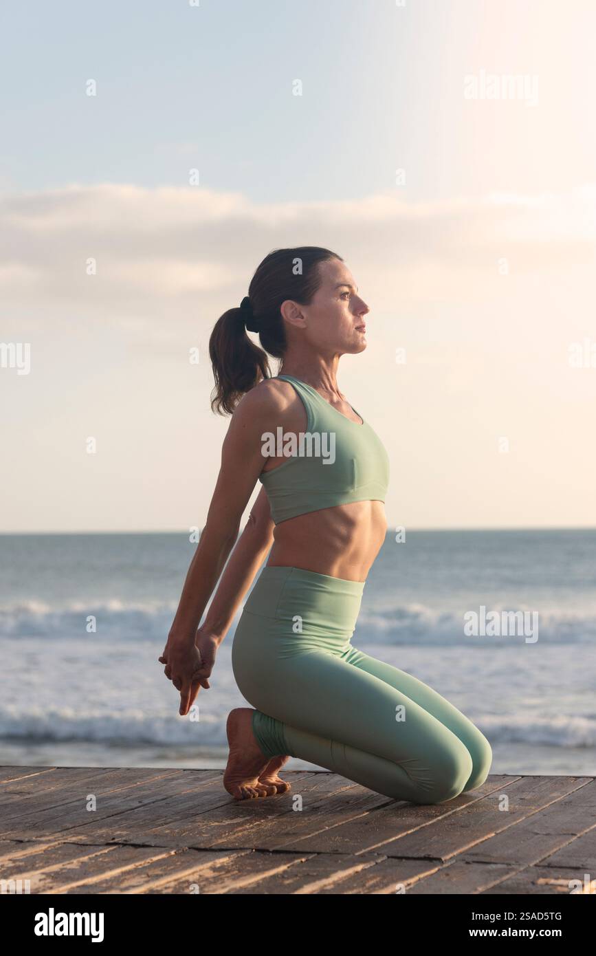 Sportliche Frau, die am Meer auf einem Steg sitzt und Yoga kniend übt, während sie mit den Armen hinter dem Rücken eine Bruststreckung macht Stockfoto