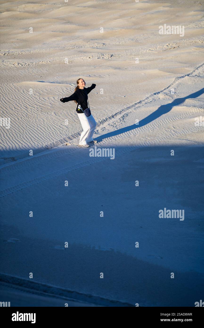 Eine fröhliche Frau in schwarzem Pullover und weißer Hose tanzt auf Sanddünen unter weichem Licht, ihre Bewegungen werfen dynamische Schatten auf das strukturierte Sandl Stockfoto