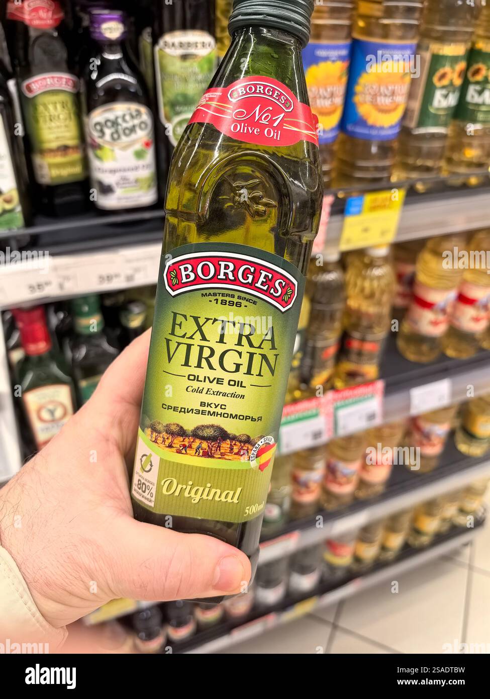 Bobruisk, Weißrussland - 24. Januar 2025: Eine Hand hält eine Flasche natives Olivenöl extra in der Hand, umgeben von einer Vielzahl von bunten Ölen in den Lebensmittelregalen. Stockfoto