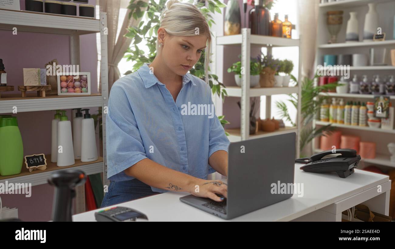 Blonde Frau, die am Laptop arbeitet, im Wohnkultur-Geschäft mit Regalen mit dekorativen Artikeln und Toilettenartikeln im Hintergrund Stockfoto