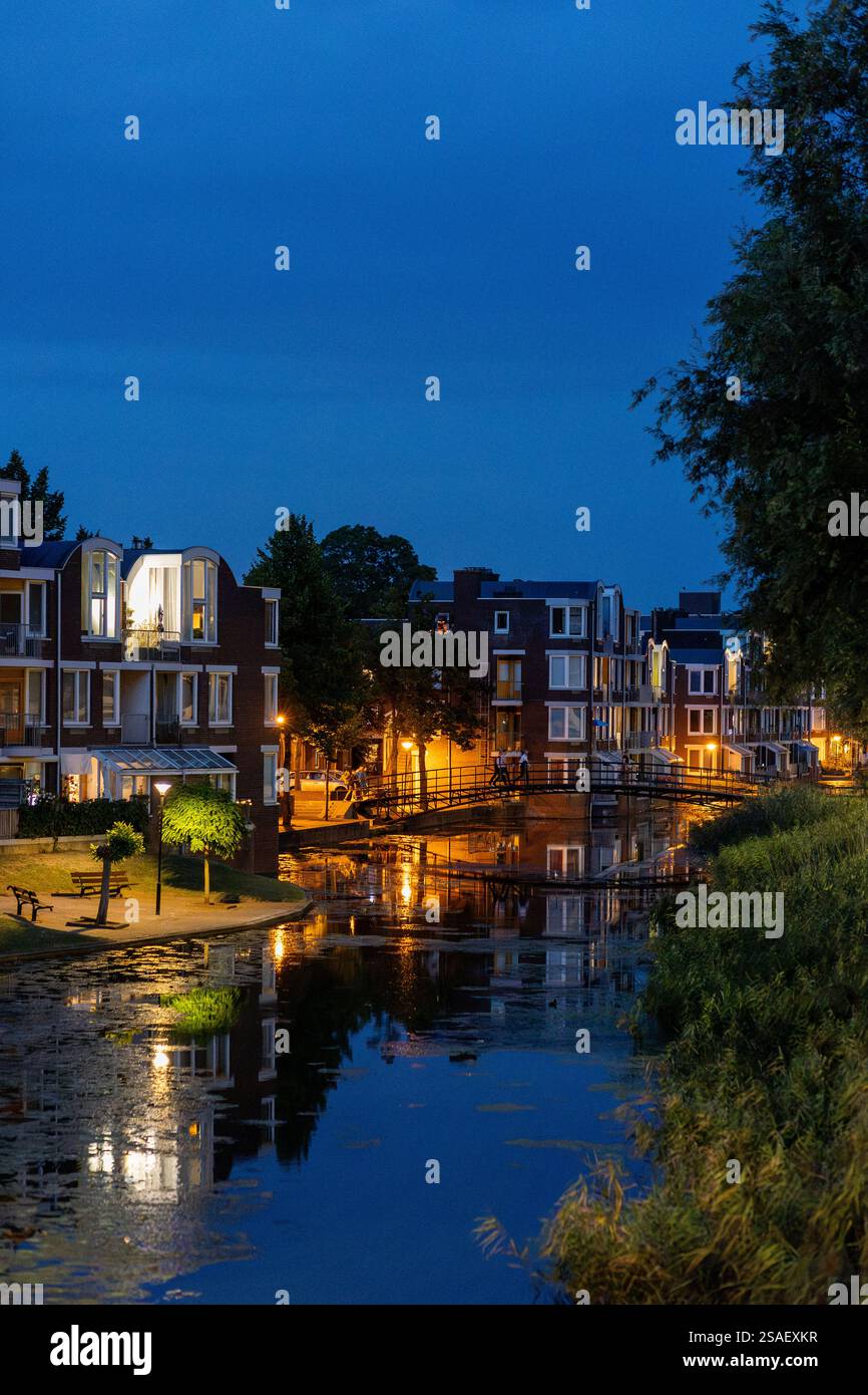 Eine ruhige Nachtszene eines modernen europäischen Viertels mit beleuchteten Gebäuden, die sich im ruhigen Kanalwasser spiegeln. Eine kleine Brücke verbindet das Haus Stockfoto