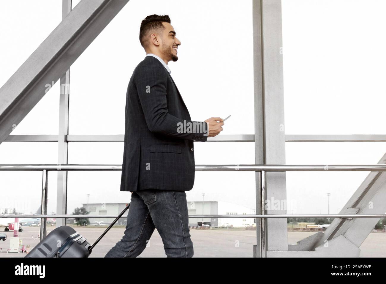 Junger Geschäftsmann aus dem Nahen Osten mit Koffer und Smartphone in den Händen, der auf dem Flughafen unterwegs ist Stockfoto
