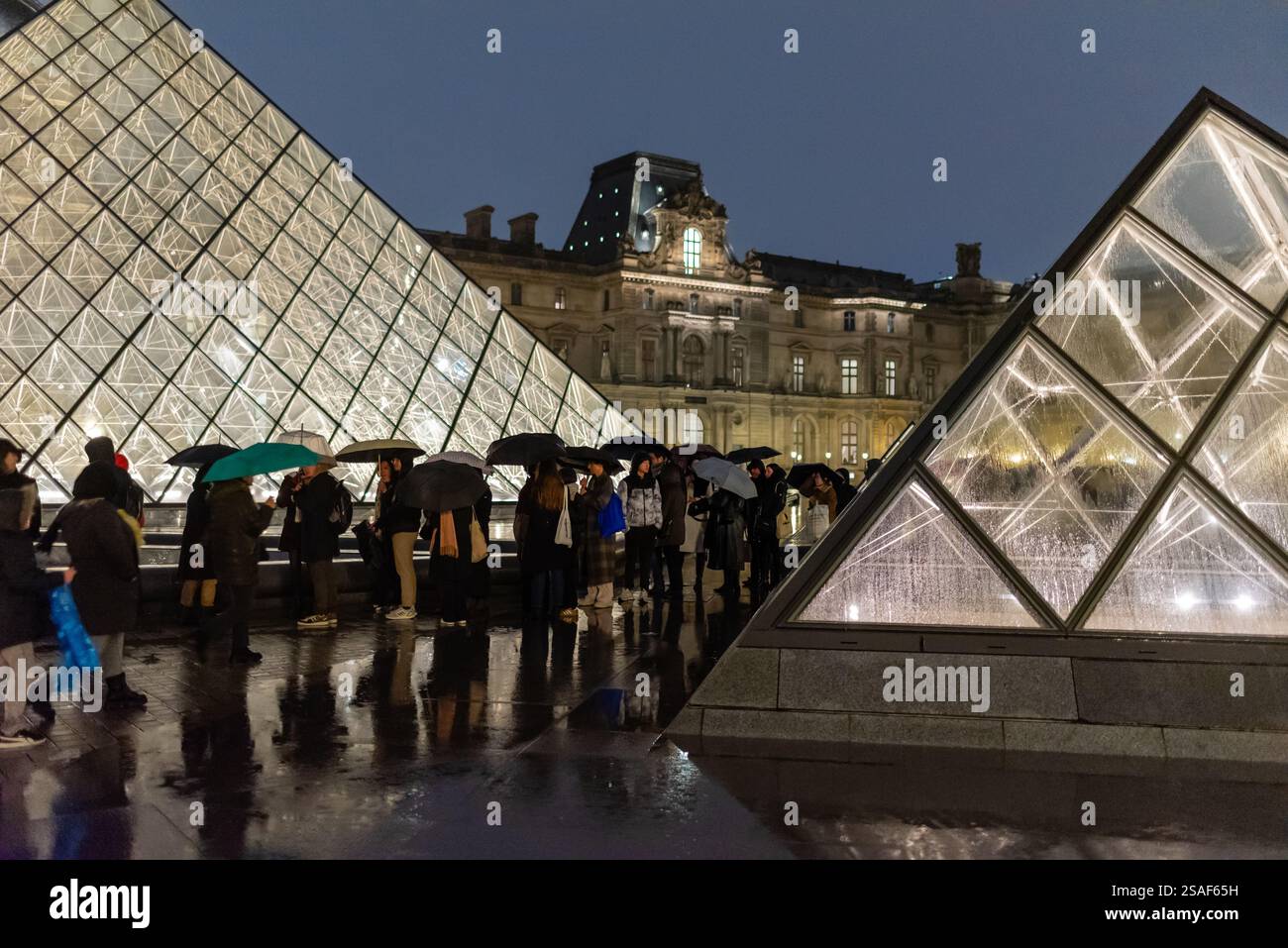 Die Besucher stehen mit Regenschirmen zwischen den Glaspyramiden des Louvre und warten auf den Eintritt in das Museum in Paris, Frankreich. Stockfoto