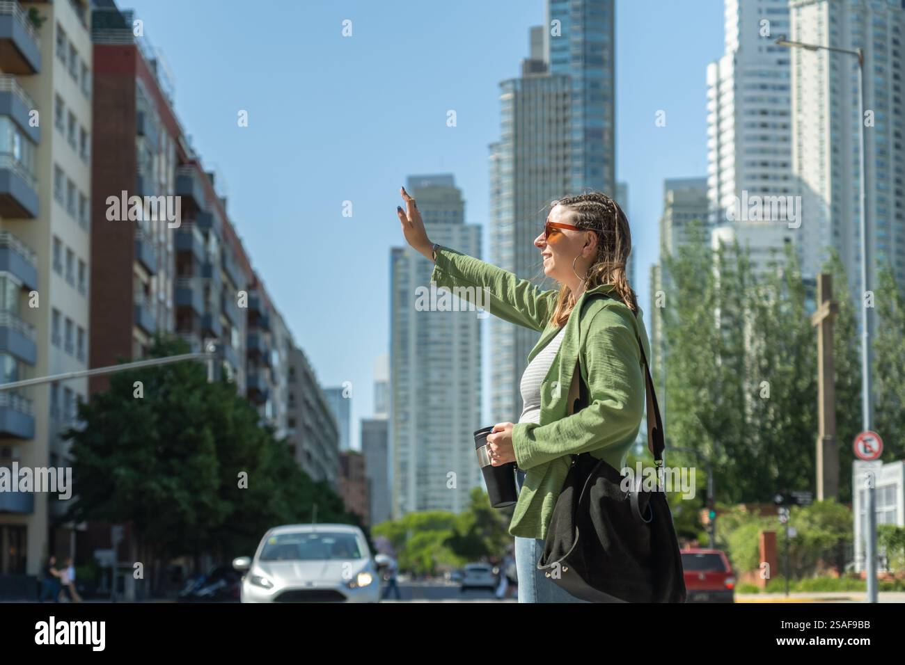 Geschäftsfrau, die an einem sonnigen Tag in buenos aires ein Taxi hält und eine wiederverwendbare Kaffeetasse hält, während sie durch die geschäftigen Straßen der Stadt mit modernem Gebäude navigiert Stockfoto