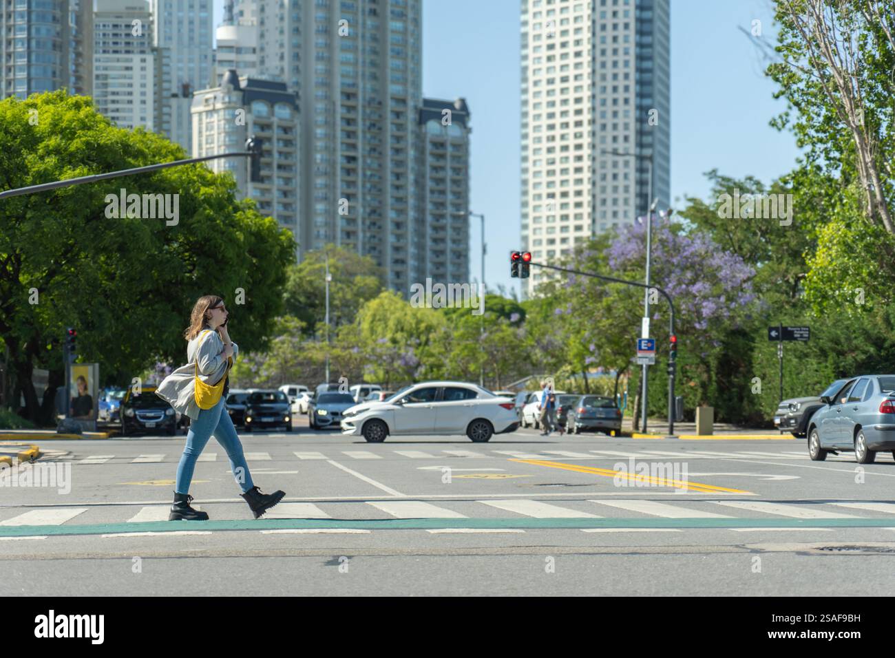 Junge Frau überquert die Straße auf Fußgängerübergängen in der Stadt, spricht mit dem Handy, während Autos auf der Straße und in der Skyscr fahren Stockfoto