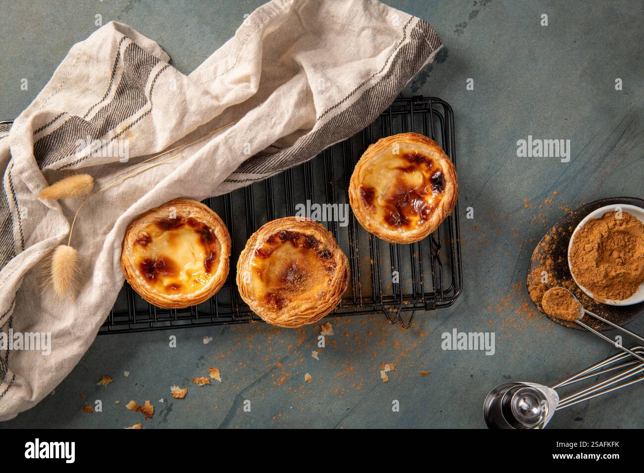 Drei frisch gebackene portugiesische Pudding-Torten auf einem Kühlregal mit einem Leinentuch und Zimt, die zum Besprühen bereit sind Stockfoto