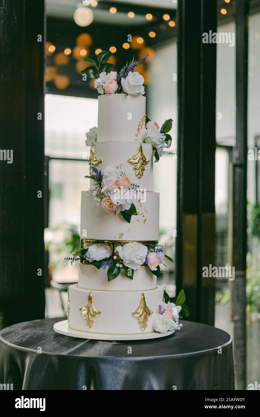 Eine wunderschön gearbeitete fünfstöckige Hochzeitstorte steht elegant auf einem runden Tisch, dekoriert mit frischen Blumen und goldenen Akzenten, in einem modernen Veranstaltungsort für Stockfoto