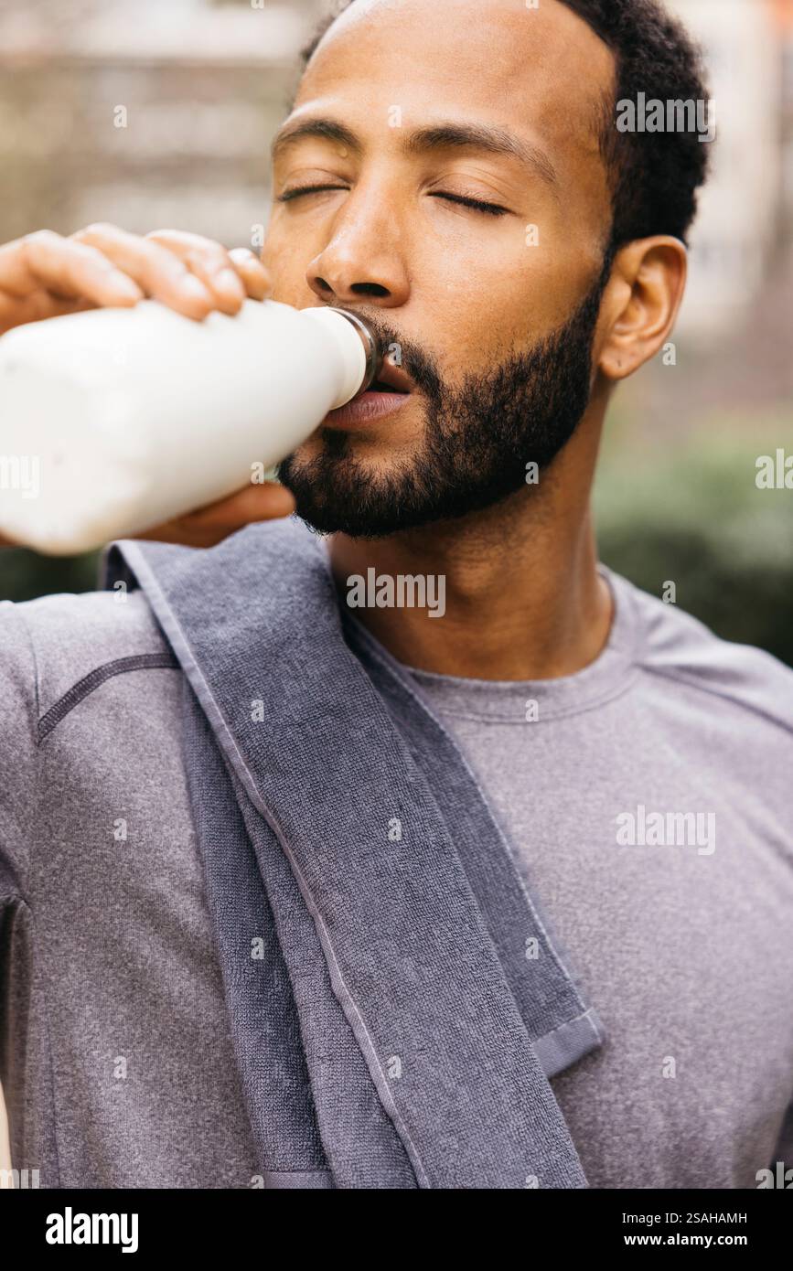 Ein sportlicher Mann erfrischt sich draußen, hält ein Handtuch und trinkt aus einer Flasche. Stockfoto
