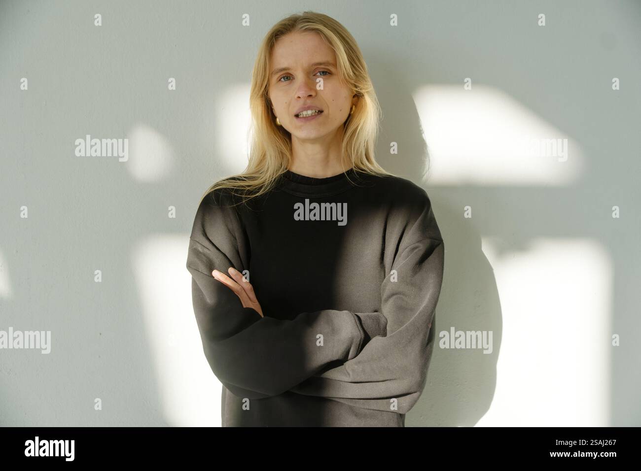 Eine junge, selbstbewusste Frau mit einer natürlichen, strahlenden Aura steht mit überkreuzten Armen vor einer sonnendurchfluteten Wand. Ihr ruhiger Ausdruck und ihr sanfter Blick strahlen Wärme aus, autonom Stockfoto