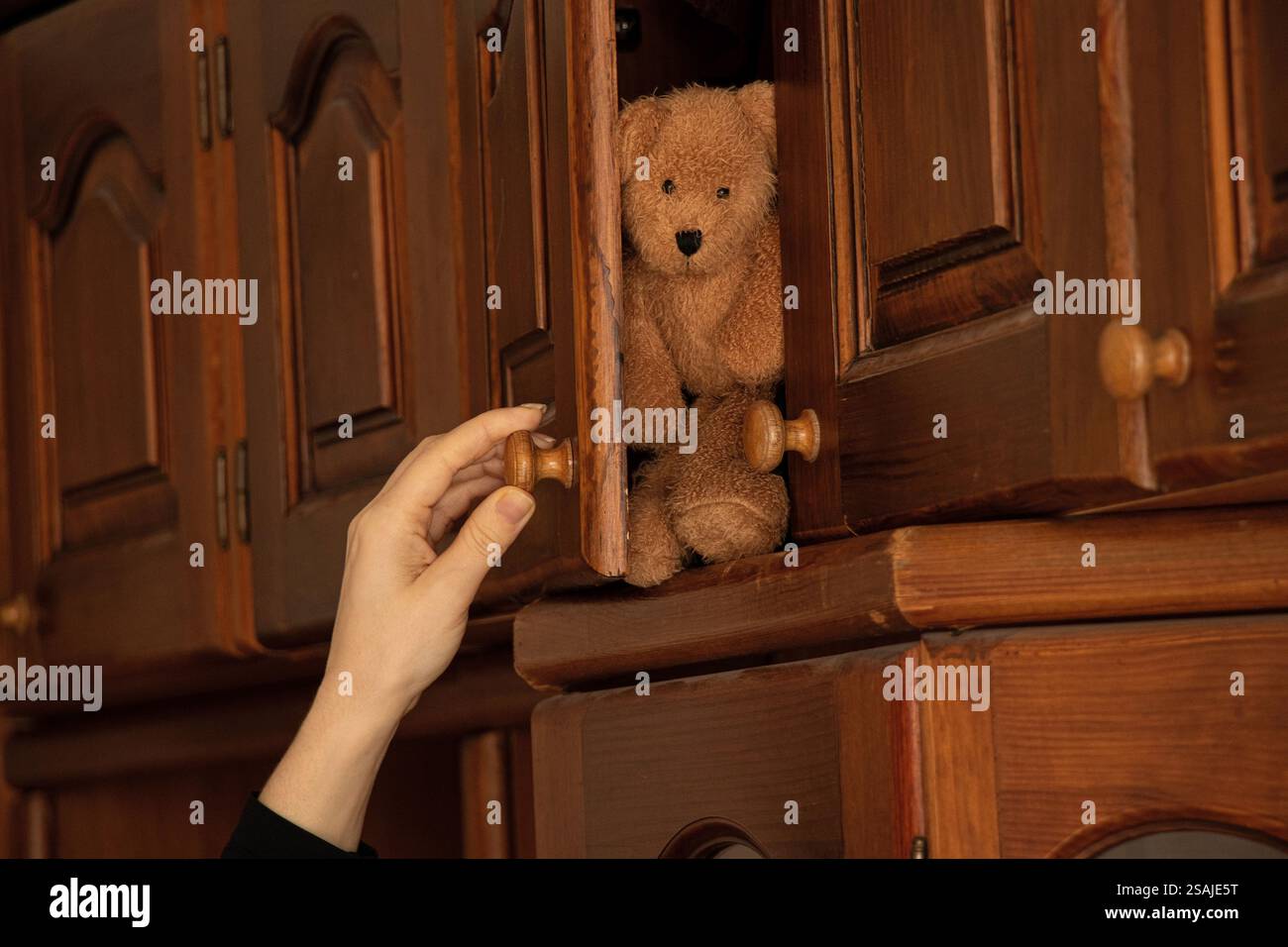 Die Hand einer Frau öffnet einen alten Holzschrank im Schlafzimmer des Hauses und auf dem Regal sitzt ein brauner Kinderteddybär. Stockfoto
