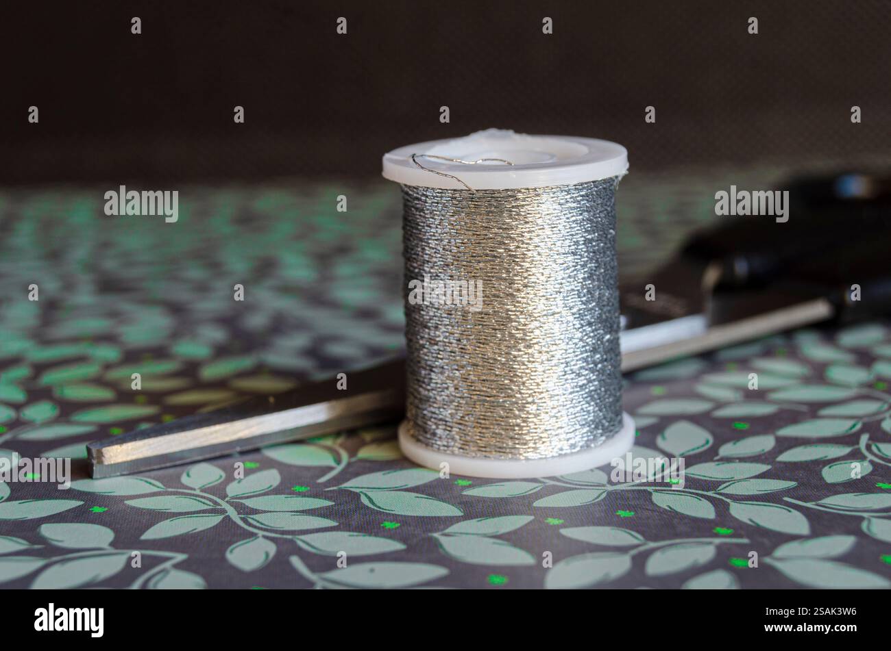 Spule aus silbernem Metallfaden und Schere auf grünem floralem Hintergrund Stockfoto