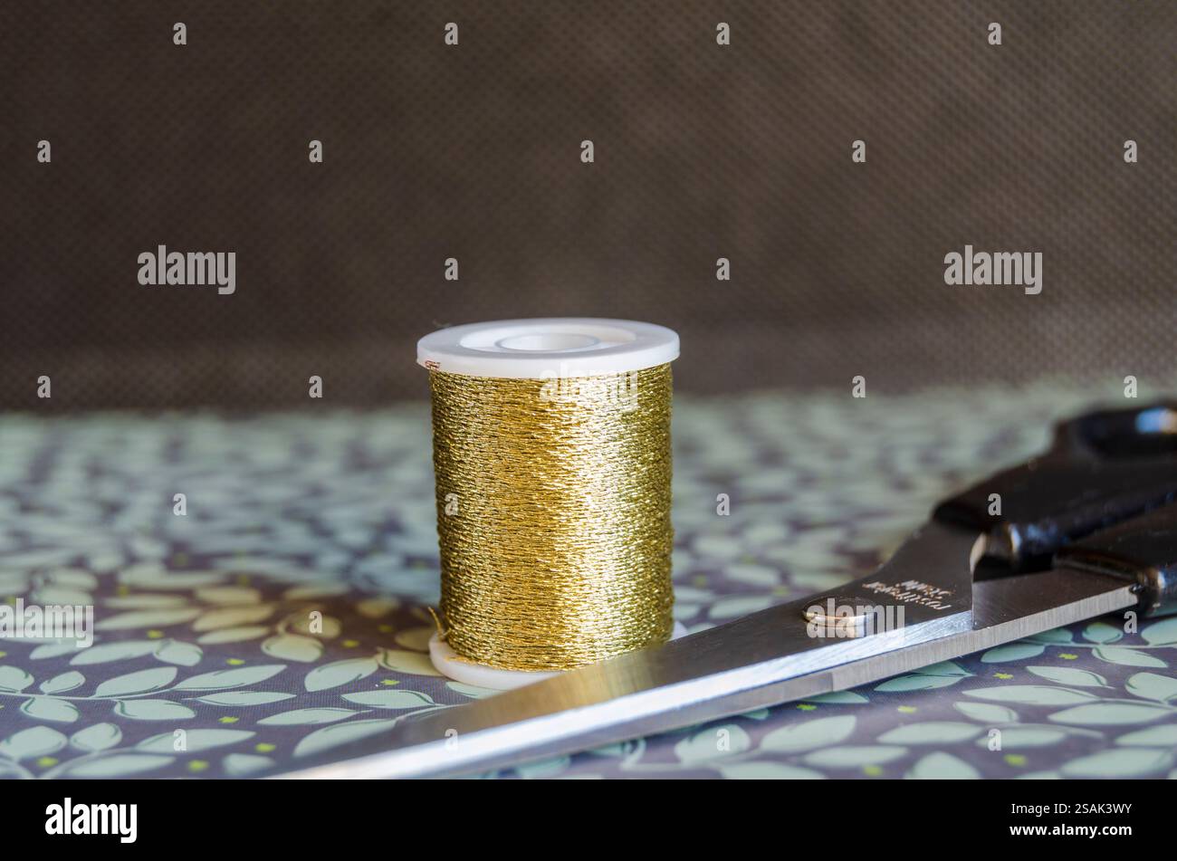 Spule aus goldenem Metallfaden und Schere auf grünem floralem Hintergrund Stockfoto