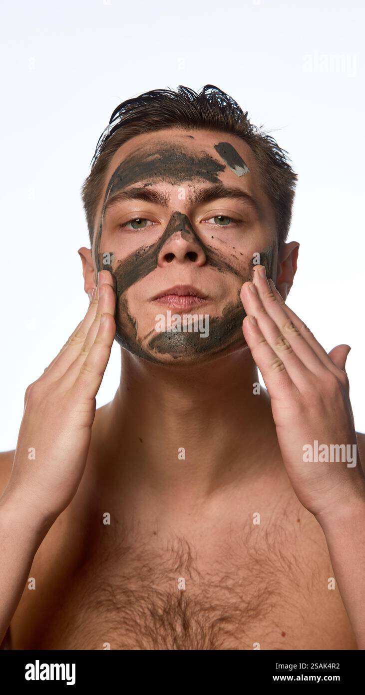 Junger Mann mit nassen, brünetten Haaren, der Hautpflegeverfahren durchführt, Nährtonmaske auf der Haut vor weißem Hintergrund aufträgt. Stockfoto