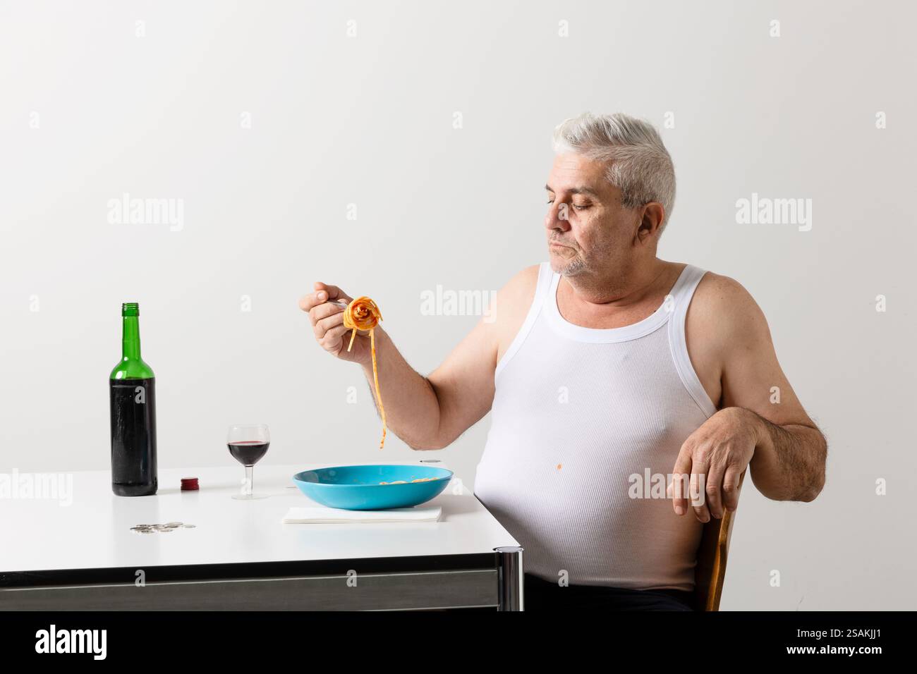 Ein Mann in seinen 60ern isst einen Teller Pasta mit Tomatensauce. Er trägt eine weiße Arbeitsweste, wird in Dreiviertelansicht gezeigt und auf dem Tisch ist eine Flasche o Stockfoto