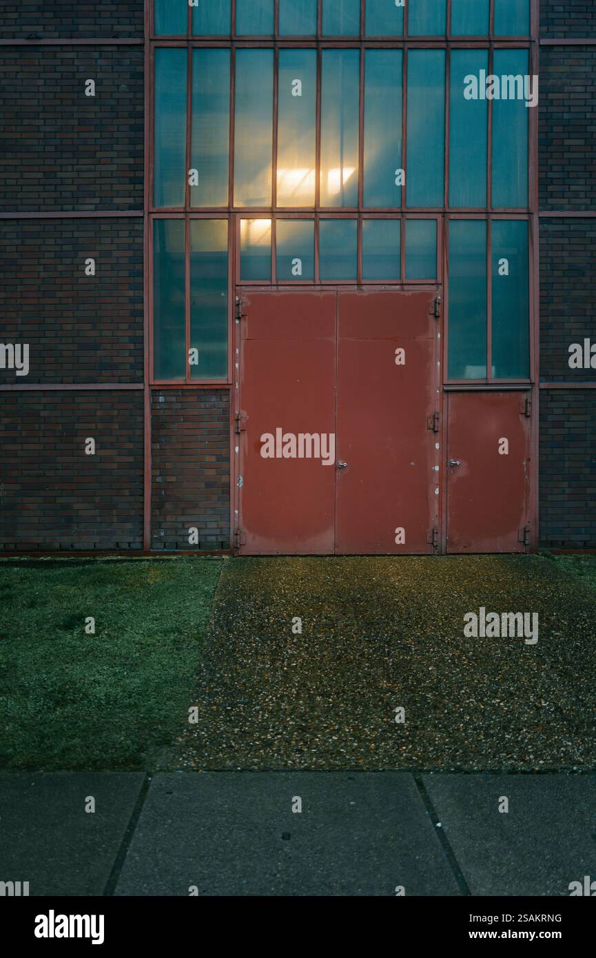 Rote Industrietür umgeben von hohen Glasfenstern und Ziegelwänden. Der Weg zur Tür ist nass und reflektiert warmes Innenlicht, Kreatin Stockfoto