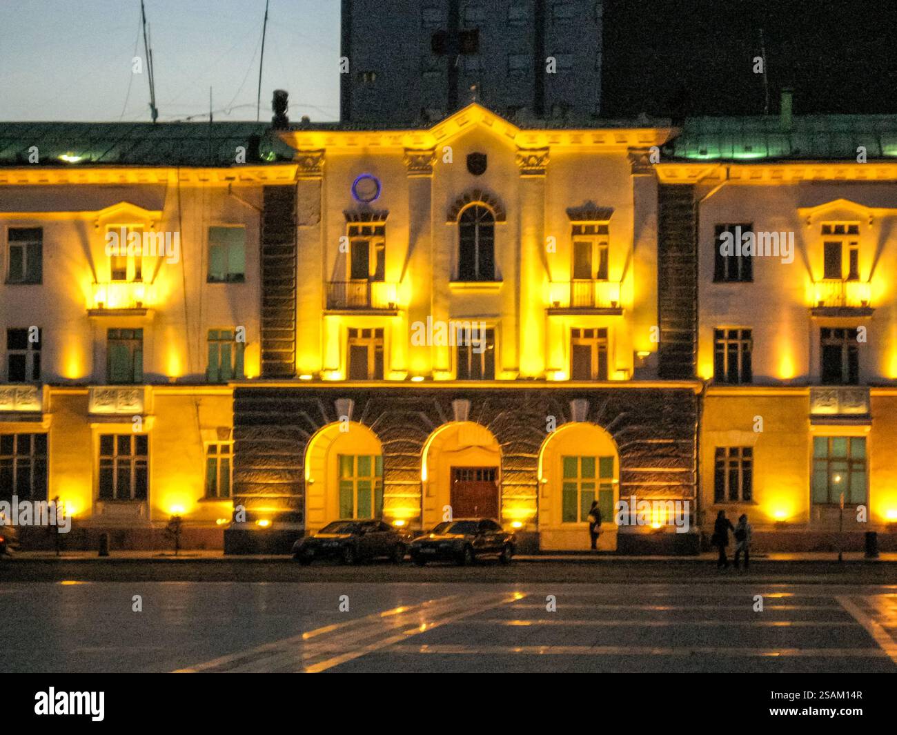 Ein großes Gebäude mit gelber Fassade wird nachts beleuchtet. Das Gebäude hat viele Fenster und einen großen Torbogen. Die Lichter am Gebäude geben ihm einen Stockfoto