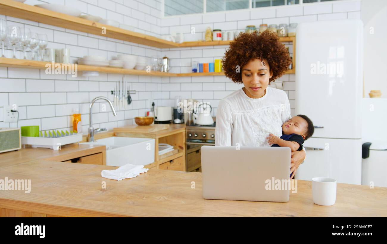 Multitasking-Mutter Hält Den Schlafenden Sohn, Während Sie In Der Küche An Einem Laptop Arbeitet Stockfoto