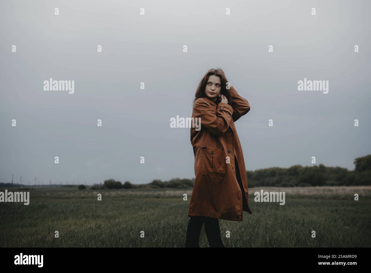 Frau in braunem Mantel auf dem Feld, bedeckter Himmel, sieht introspektiv und friedlich aus Stockfoto