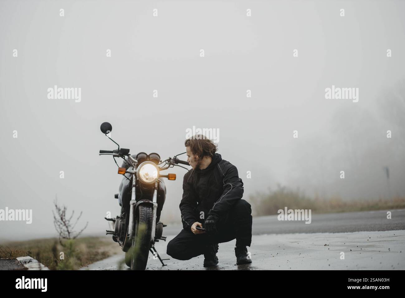 Die Person hockt sich neben einem Motorrad auf einer nebeligen Straße und schafft eine einsame und introspektive Stimmung Stockfoto