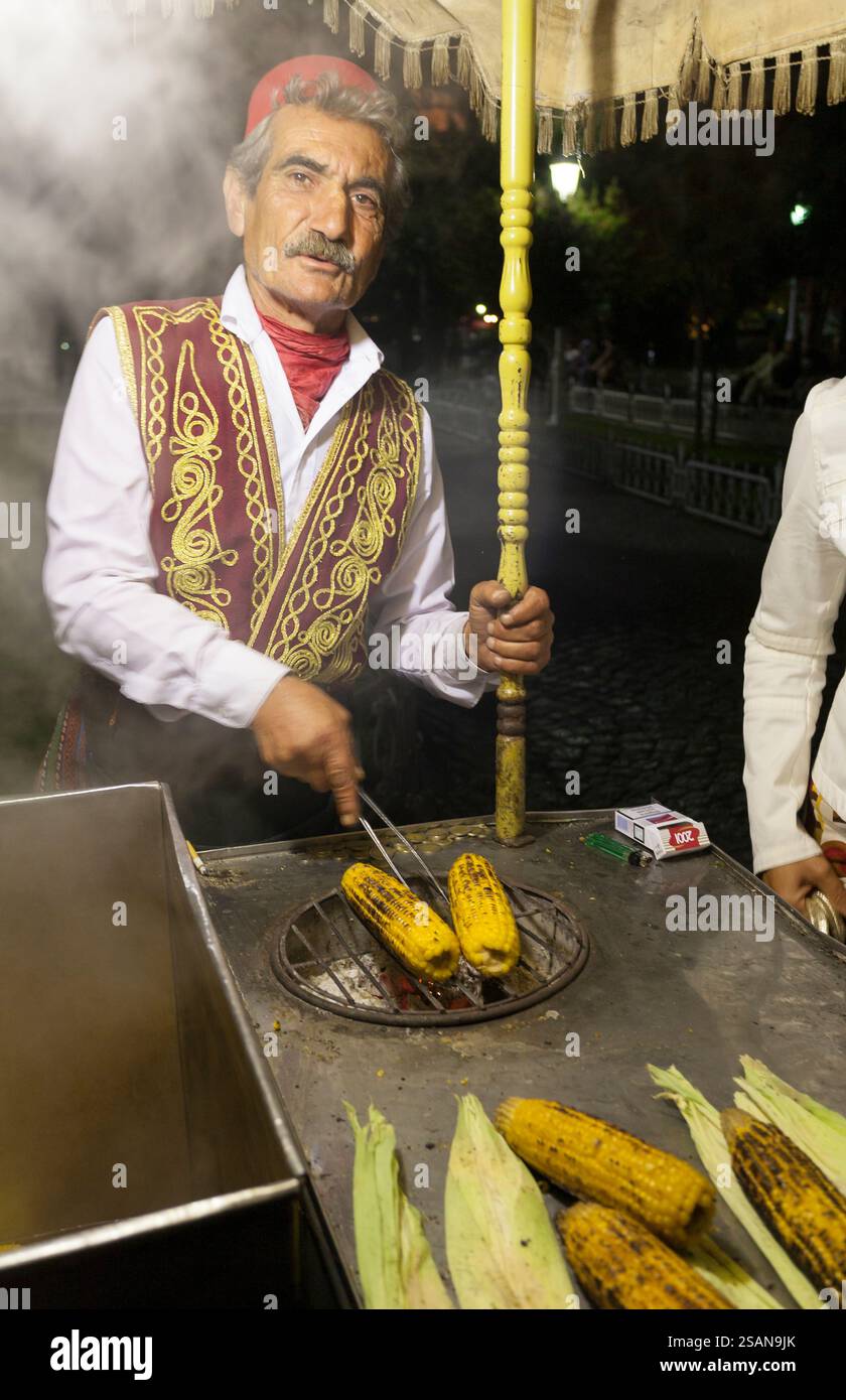 Corn Roasting Street Vendor in Sultanamet: Ein Straßenverkäufer, der in traditioneller Tracht gekleidet ist, röstet gelbe Maiskolben, um sie an hungrige Passanten zu verkaufen, nachdem das Fasten am Abend unterbrochen wurde. Stockfoto