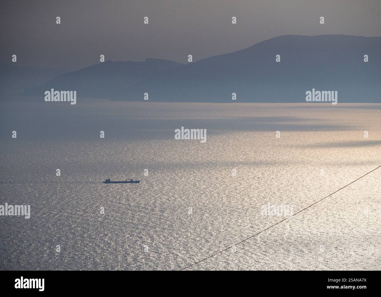 Folegandros Sonnenaufgang - Ein Frachtschiff sprüht das sonnendurchflutete Wasser: Die hohen Berge von Sikinos erheben sich aus dem Morgennebel und Nebel im Hintergrund, während die warme Sonne das ruhige Meer mit Licht betäubt. Stockfoto