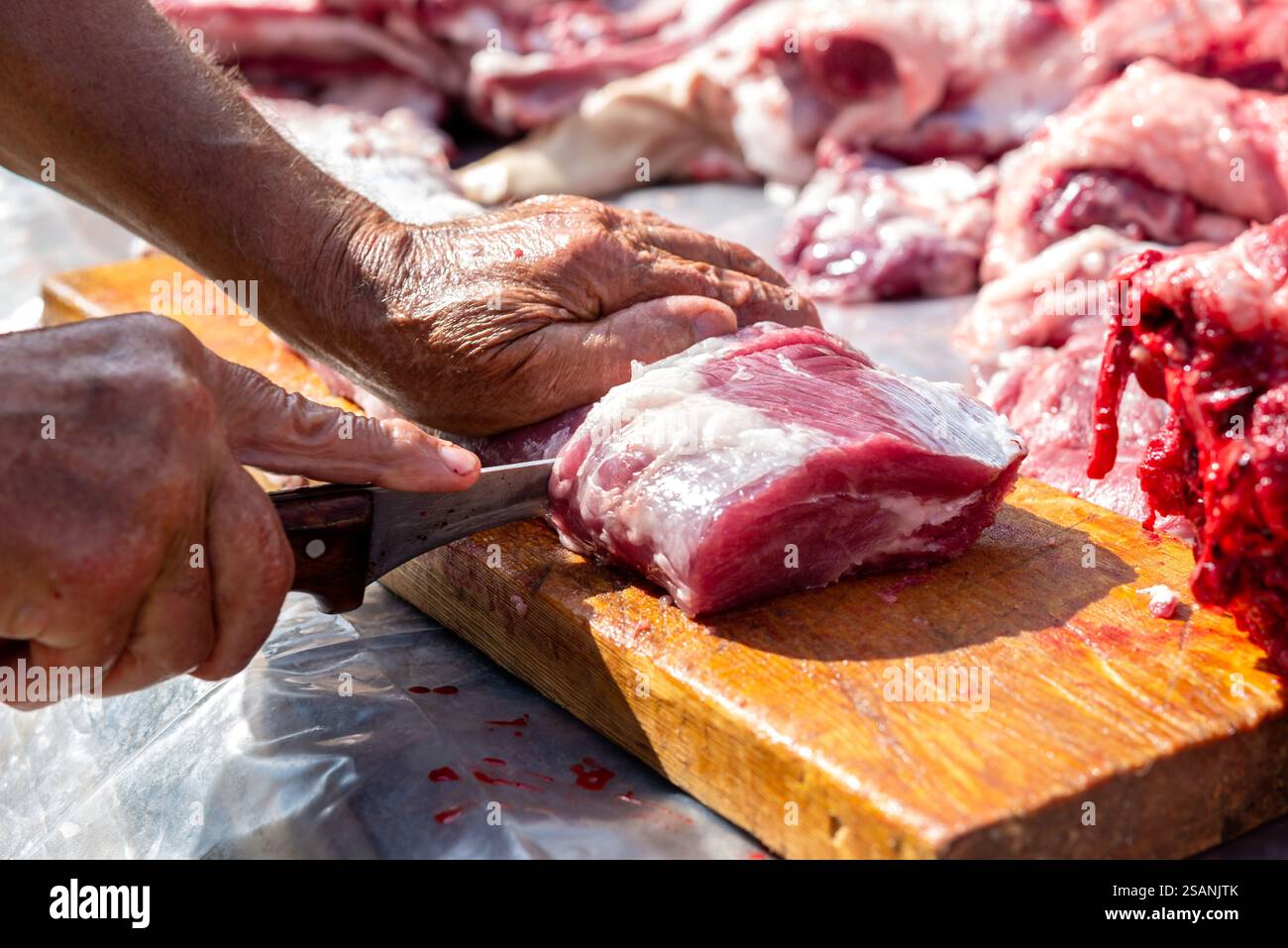 Die Hände des alten Mannes schneiden rohes Schweinefleisch mit einem Messer auf einem hölzernen Schneidebrett draußen. Stockfoto