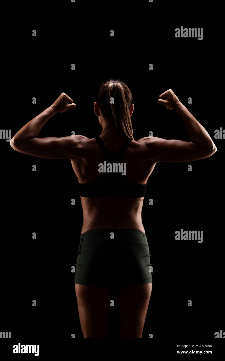 Ein kraftvolles, hintergrundbeleuchtetes Bild einer Frau in Fitnesskleidung, das Stärke und Entschlossenheit zeigt, indem sie ihre Armmuskeln vor schwarzem Hintergrund beugt. Stockfoto