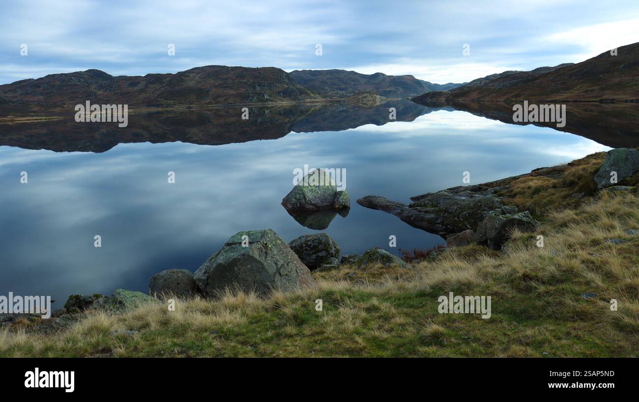 Ruhige Landschaft mit ruhigem Wasser, das tagsüber den bewölkten Himmel auf einem See vom felsigen Ufer reflektiert. Norwegen Stockfoto