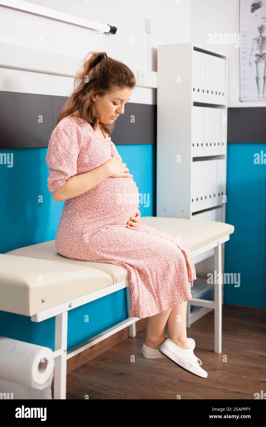 Schwangere weiße Dame sitzt allein in der Arztpraxis und wartet auf ihre Entbindungsberatung mit dem Arzt. In lässiger Kleidung streichelt die kaukasische Frau sanft ihren Schwangerschaftsbauch, während sie auf dem Krankenhausbett sitzt. Stockfoto