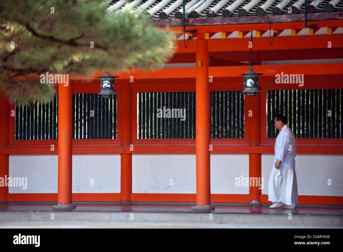 A priest dressed in white hakama pants and kosode shirt walks along a covered vermilion hallway at Heian Jingu Shinto shrine in Kyoto, Japan. Stockfoto