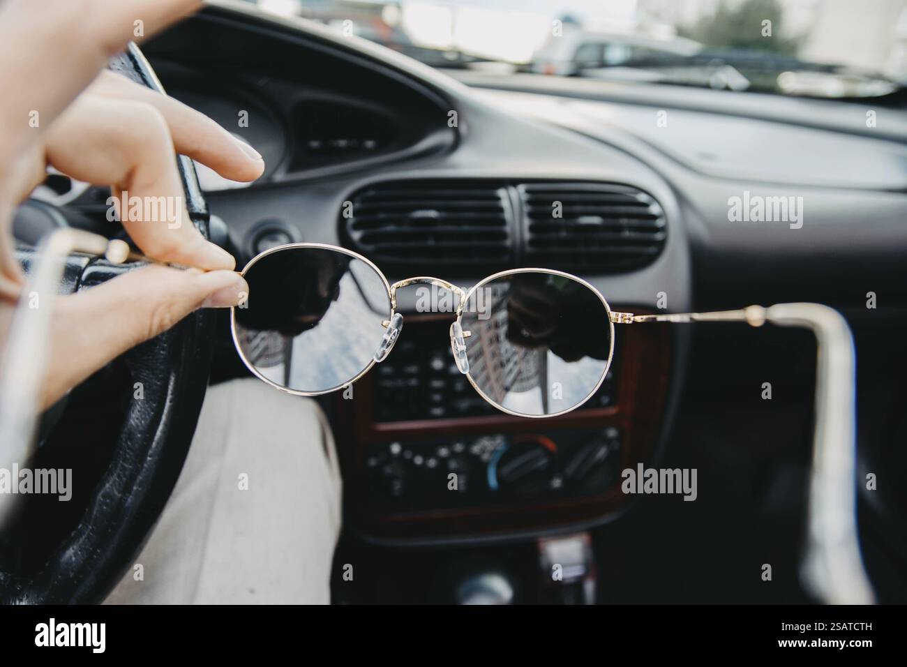 Person, die eine Sonnenbrille in einem Fahrzeug hält und eine städtische Umgebung reflektiert Stockfoto