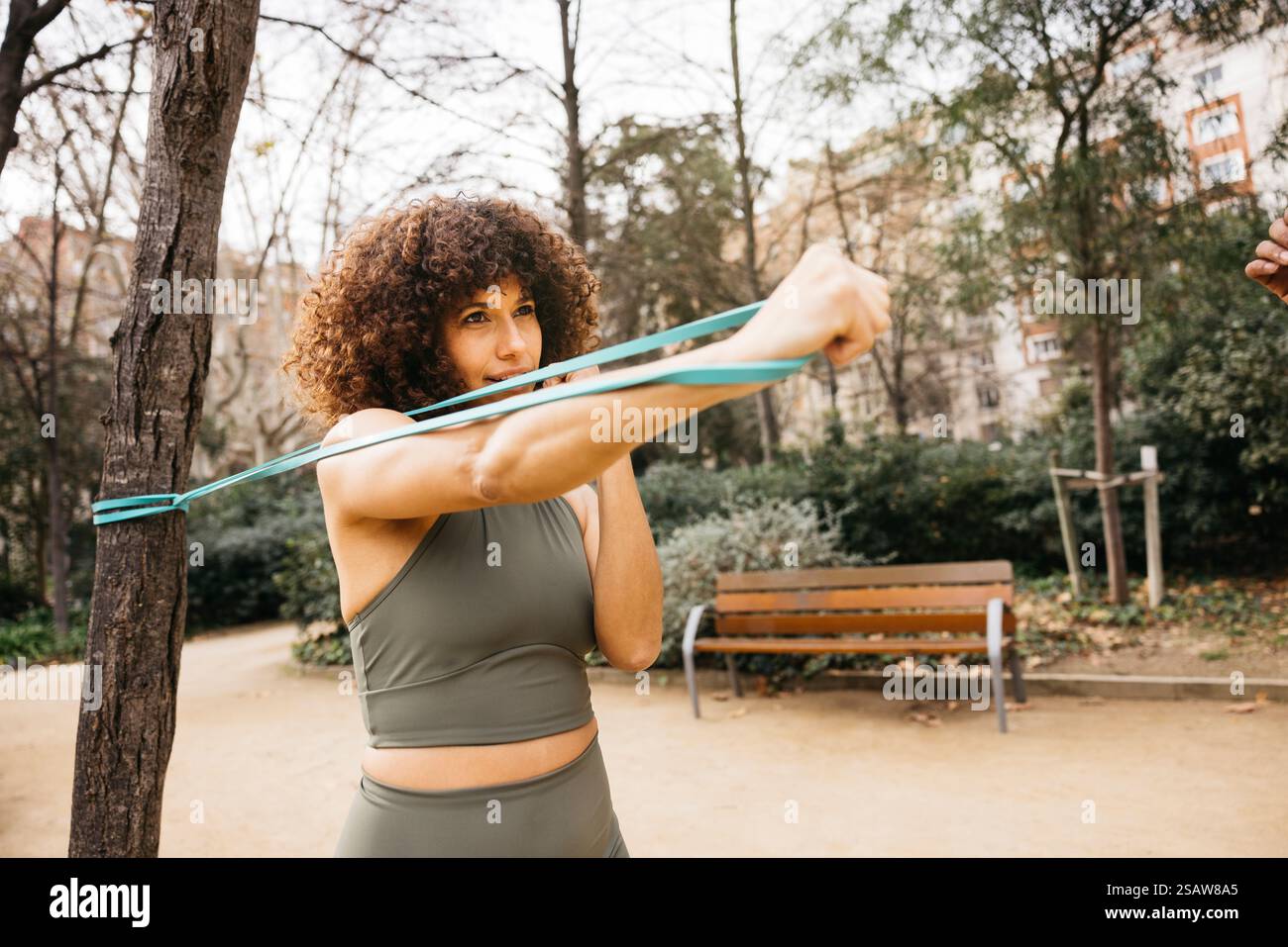 Eine Frau in Sportbekleidung führt Krafttraining mit einem elastischen Widerstandsband in einer von Grün und Bänken umgebenen Parklandschaft durch. Stockfoto