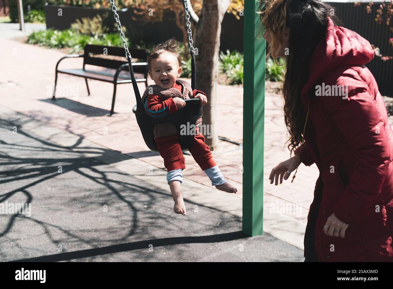 Mutter schiebt fröhliches Baby auf die Schaukel und lacht in der Wintersonne Stockfoto