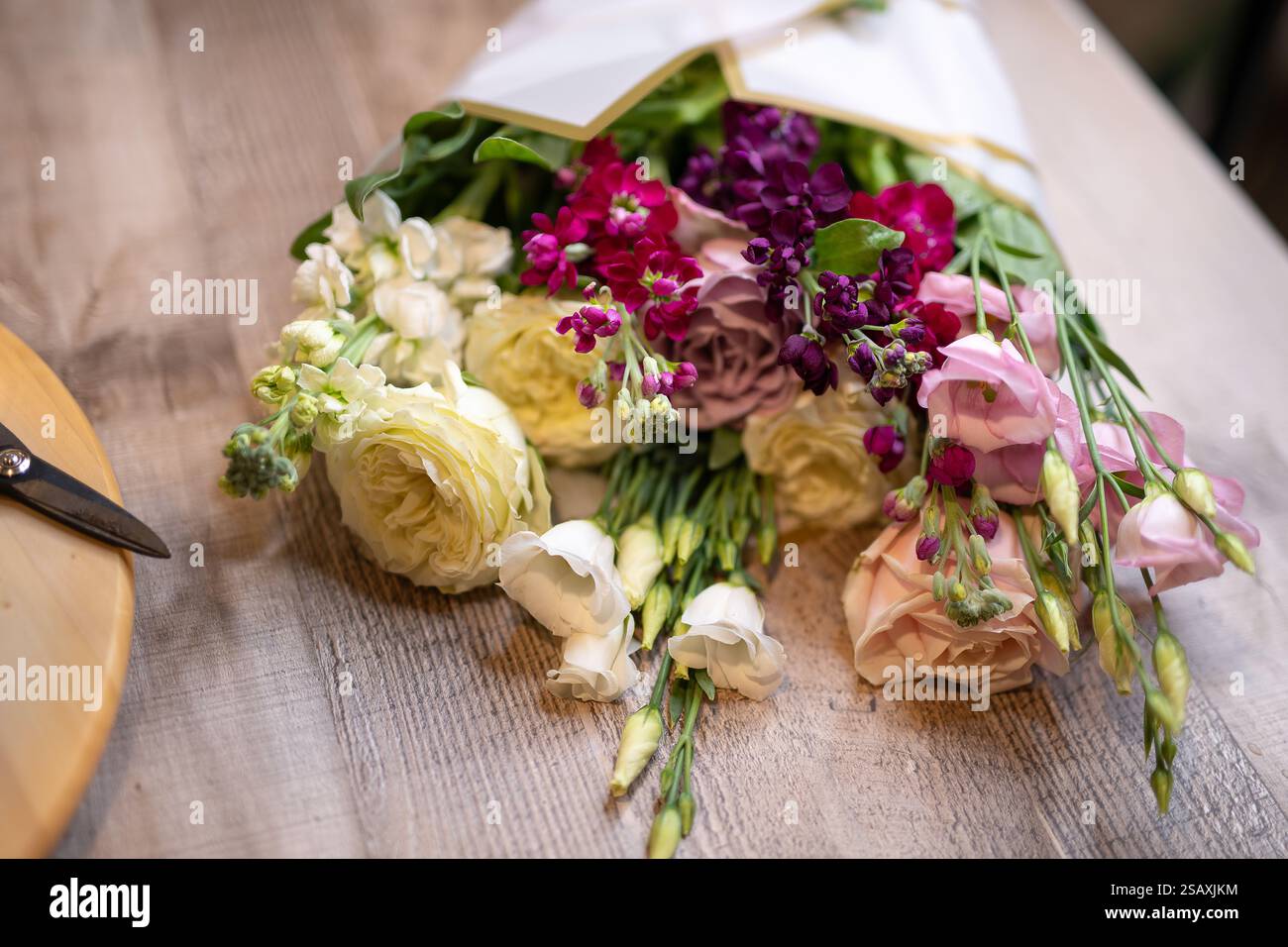Schöne Auswahl an lebendigen Blumen, die in Papier gewickelt sind, mit einer gelben Schere in der Nähe auf einer Holzoberfläche, die Kreativität und Vorbereitung veranschaulicht Stockfoto