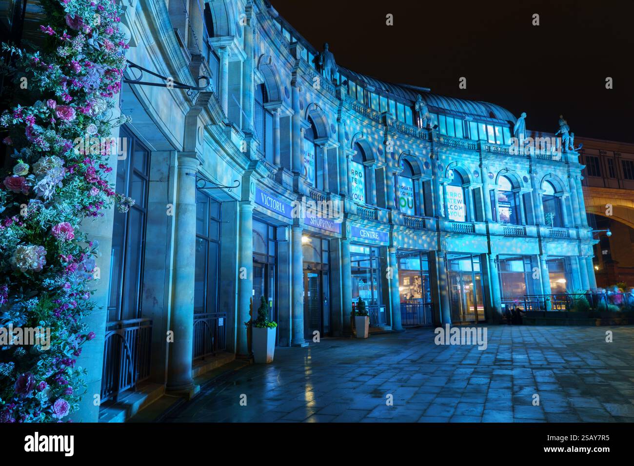 Atemberaubende nächtliche Szene mit geschwungenem Gebäude beleuchtet. Helle blaue Beleuchtung und üppige Blumendekorationen unterstreichen die atemberaubende Architektur. Harrogate, Großbritannien. Stockfoto