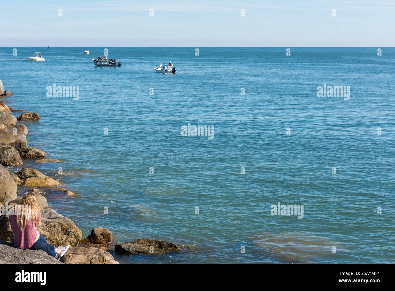 Eine Frau mit langen blonden Haaren sieht man von hinten auf einem Felssteg mit Blick aufs Meer, wo Leute auf Booten fischen, Ponce Inlet, Florida. Stockfoto