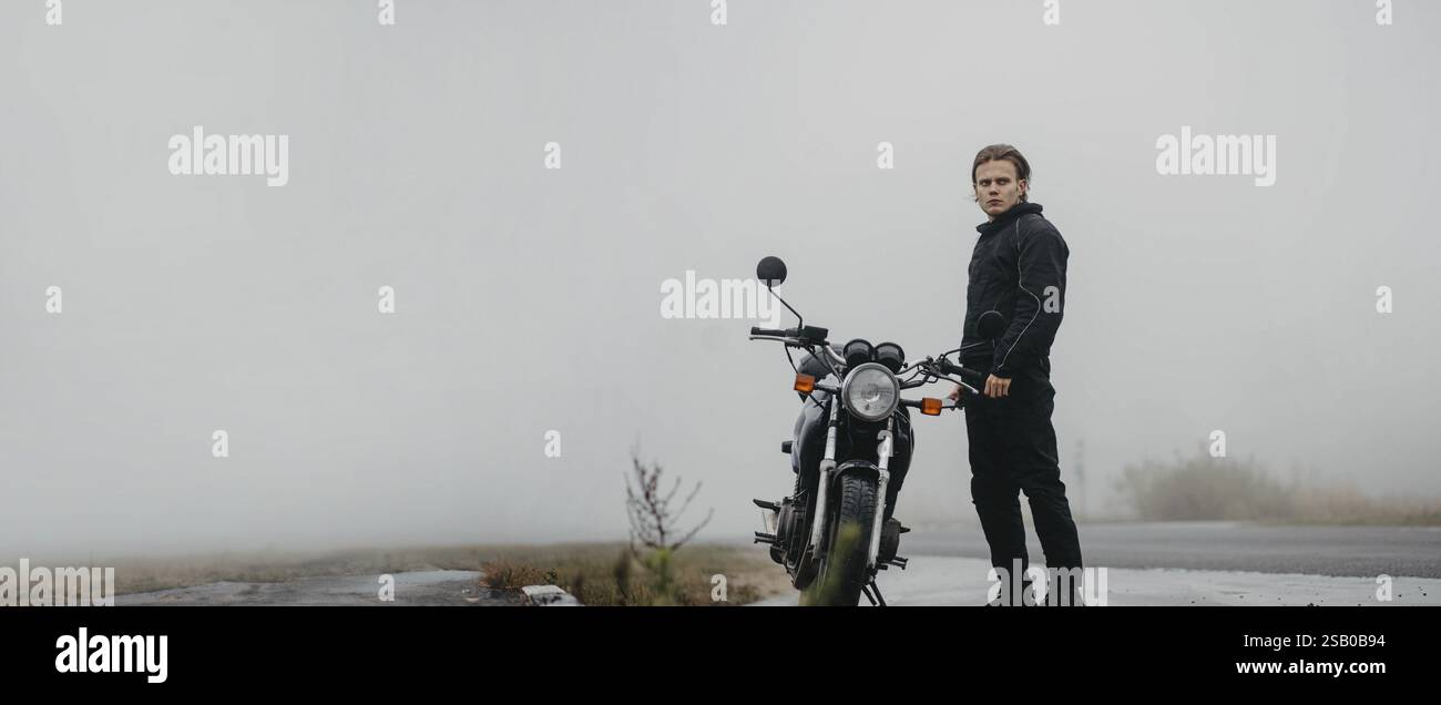 Ein junger Mann steht an einem Motorrad auf einer nebeligen Straße und blickt nach vorne Stockfoto