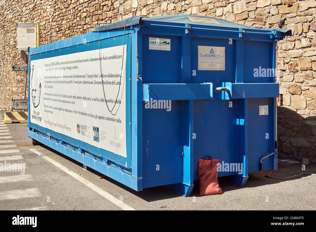 Civitavecchia. Italien - 31. Januar 2025: Großer blauer Abfallcontainer in städtischer Umgebung vor einer Steinmauer. Der Behälter hat verschiedene Etiketten und sig Stockfoto