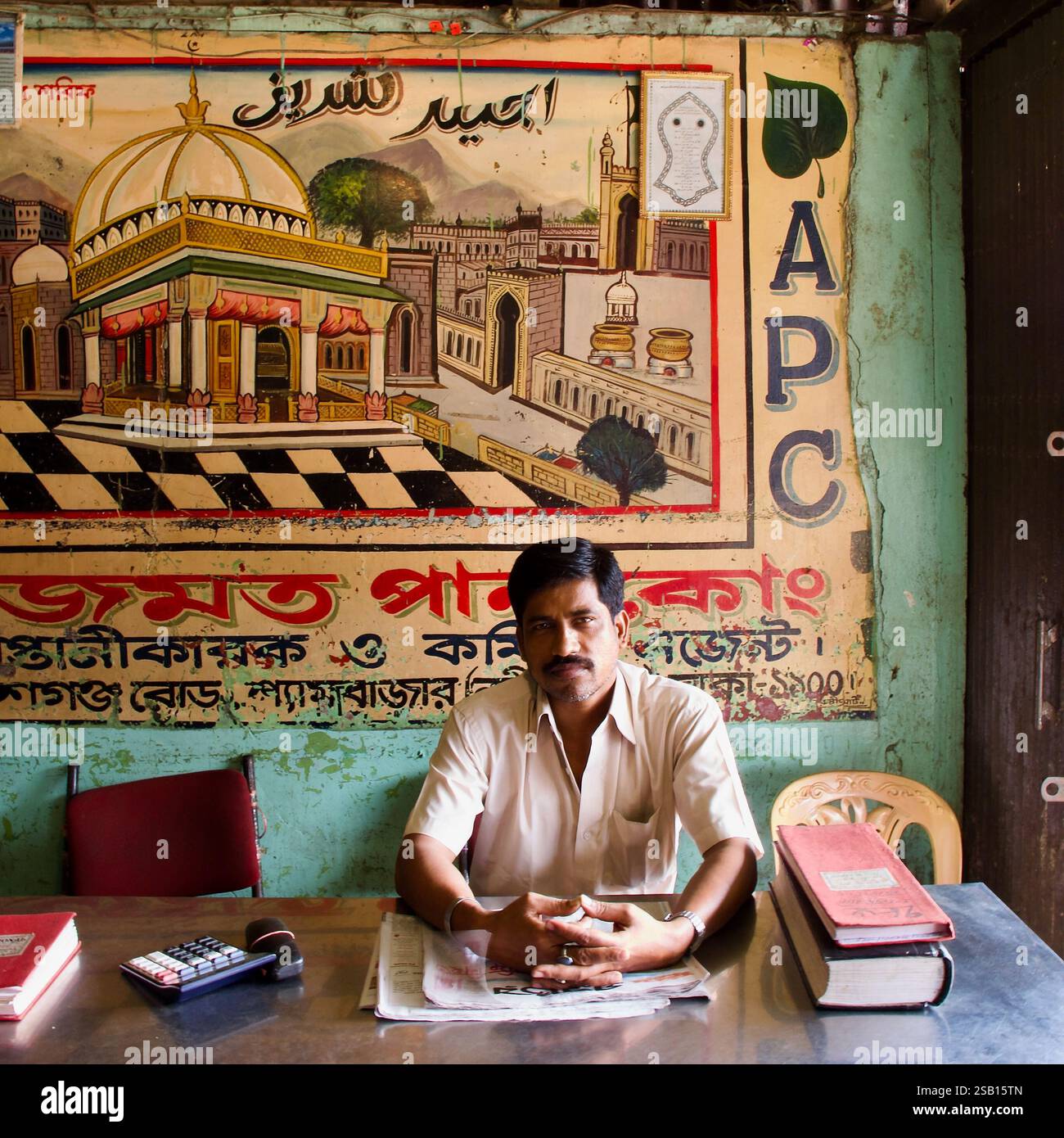 Ein einheimischer Geschäftsmann sitzt an seinem Schreibtisch in Dhaka, Bangladesch, unter einem handgemalten islamischen Wandgemälde, das Glauben und Handel im Alltag vereint. Stockfoto