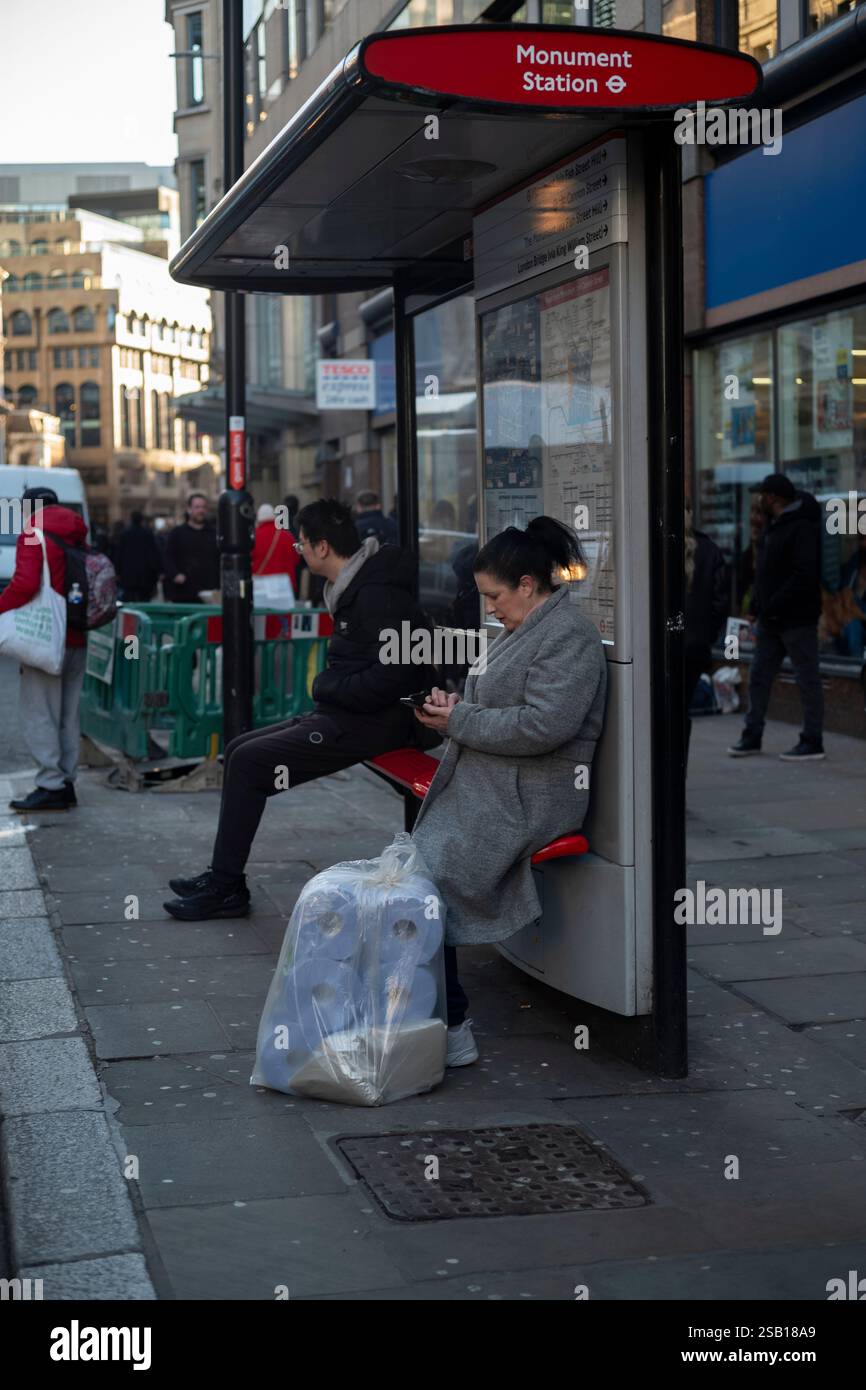 Eine Dame sitzt an einer Bushaltestelle entlang der Eastcheap, mit einer Tasche mit Rollen blauem laminiertem Papier, in der City of London, England, Großbritannien Stockfoto