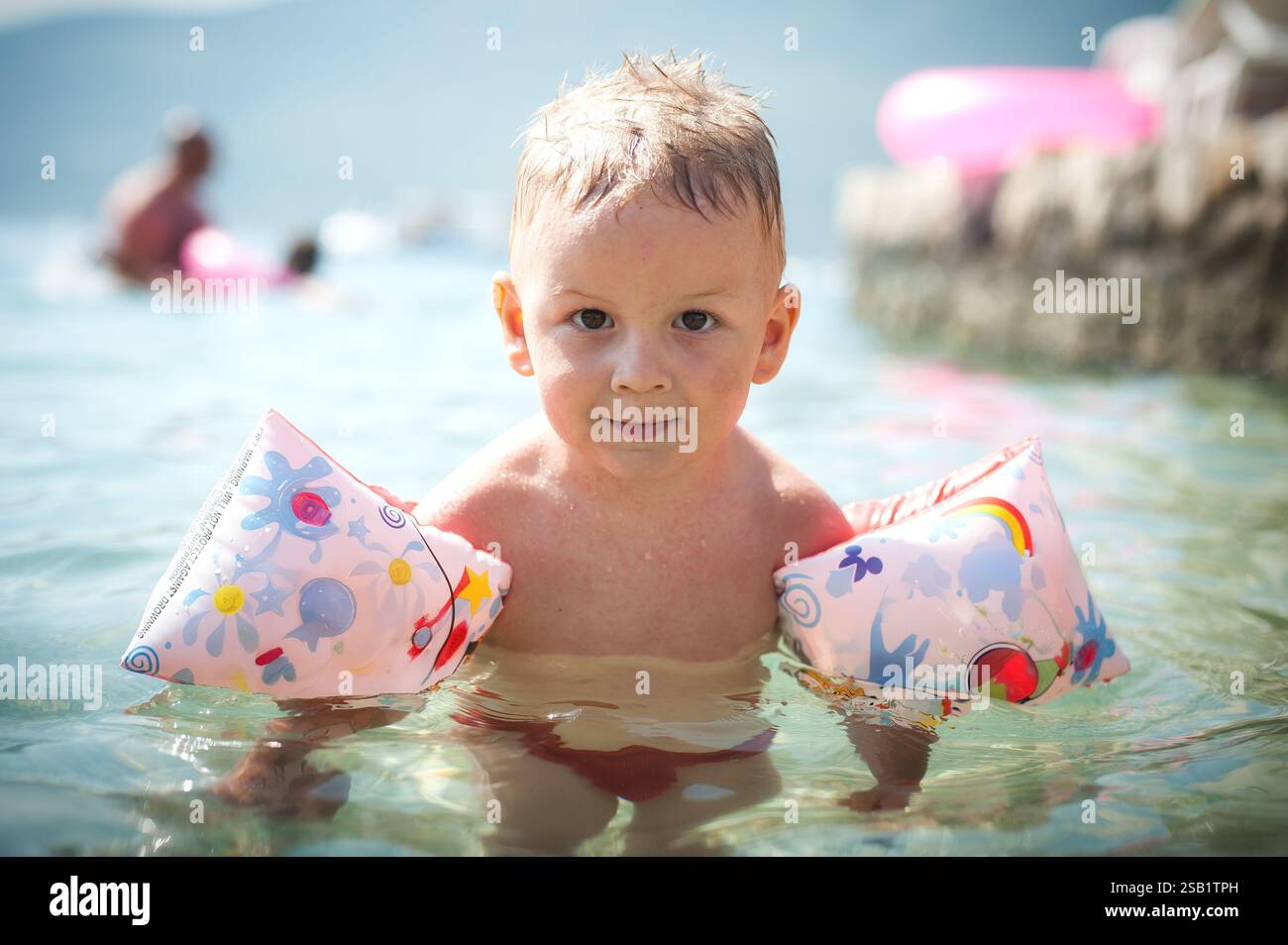 Helle Sonne reflektiert das klare Wasser, während ein kleines Kind in flachen Gewässern spielt und bunten aufblasbaren Armbändern einen unbeschwerten Sommertag genießt Stockfoto