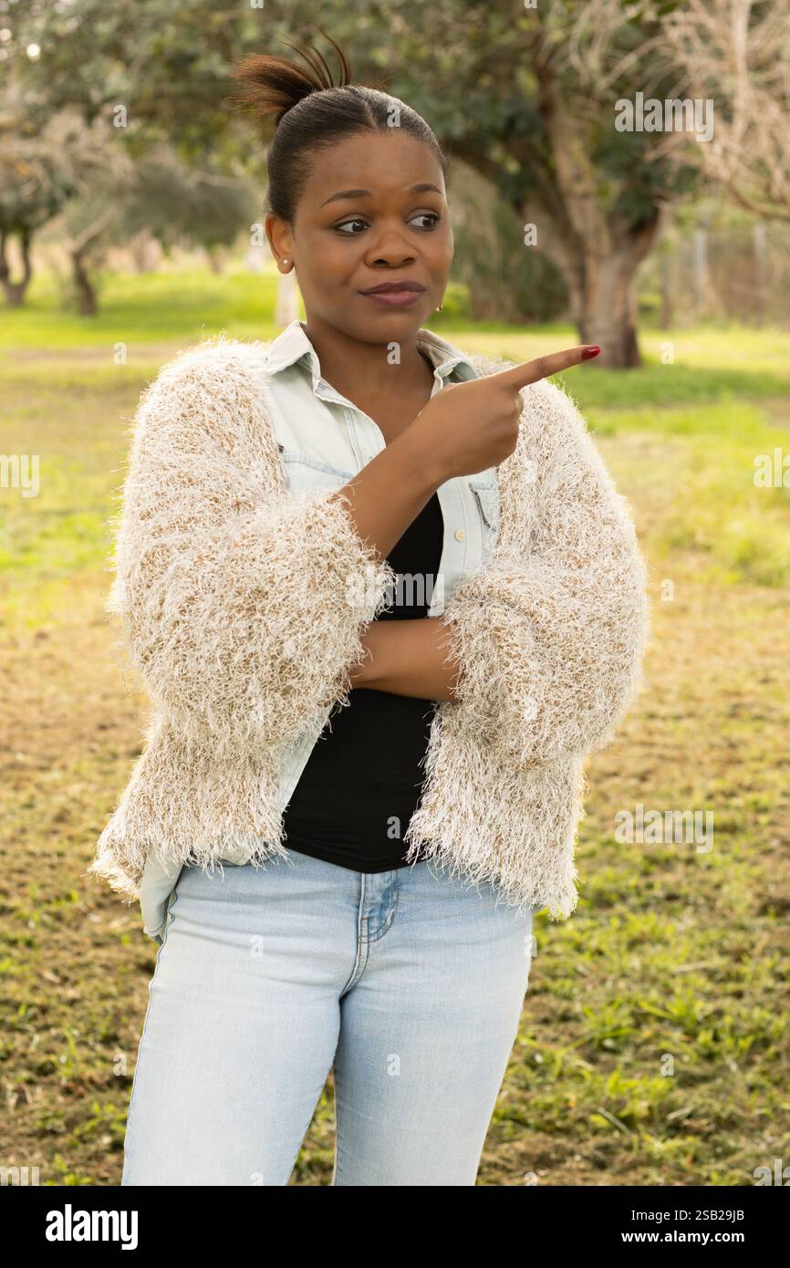 Eine schwarze Frau zeigt mit skeptischem Gesichtsausdruck zur Seite, während sie in einer natürlichen Umgebung steht. Stockfoto