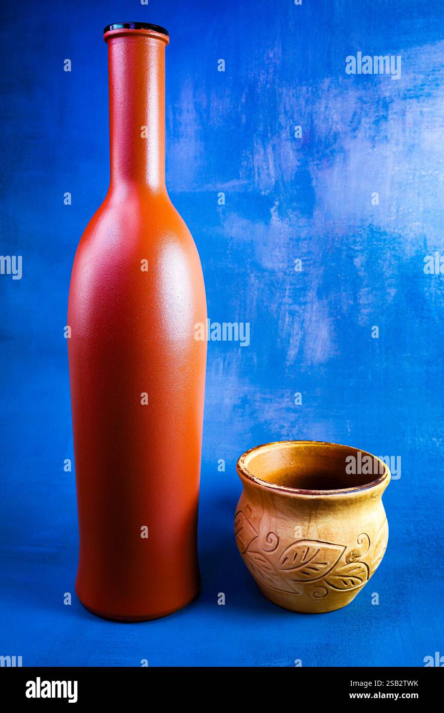 Undurchsichtige braune Glasflasche mit engem Hals und handgemachter Tonbecher auf einem hellen blauen Hintergrund Stockfoto