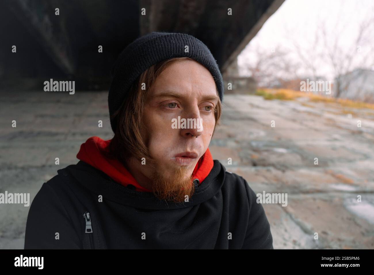 Ein junger Mann mit einem nachdenklichen Ausdruck sitzt leise unter einer Brücke, verloren in der Besinnung. Die kühle Luft deutet auf den nahenden Winter hin. Stockfoto