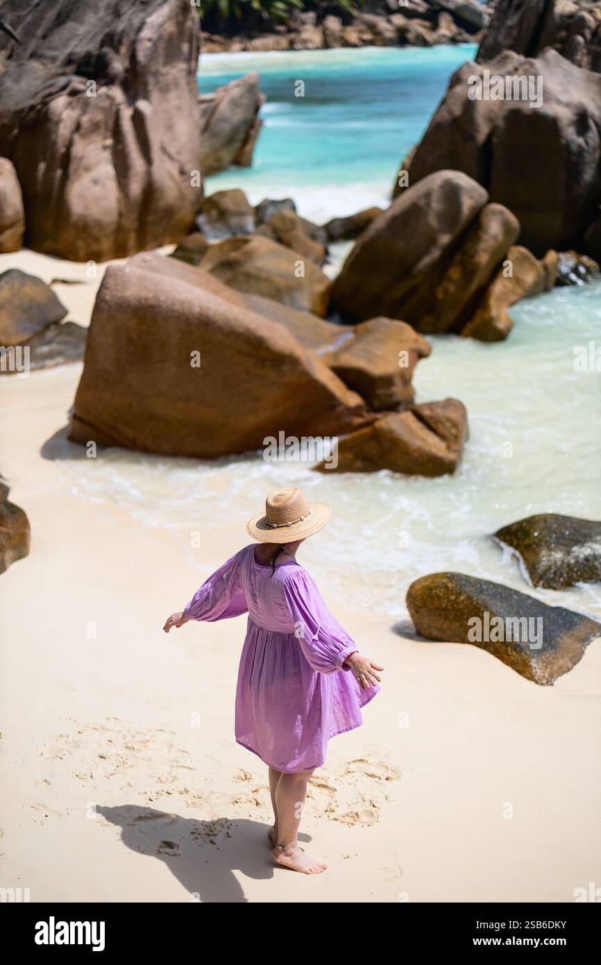 Eine Frau in rosa Kleid und Strohhut spaziert entlang eines Sandstrandes in der Nähe großer Granitfelsen mit türkisfarbenem Wasser im Hintergrund Stockfoto