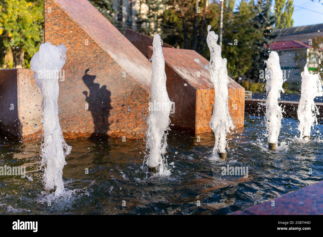 Sanfte Wasserstrahlen brechen aus Steinbauten aus und schaffen eine lebhafte Atmosphäre in einem belebten Stadtpark. Besucher genießen die erfrischenden Geräusche und Sehenswürdigkeiten Stockfoto